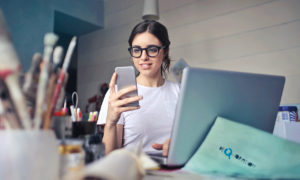 Woman sitting at a desk with a laptop looking at a smart phone